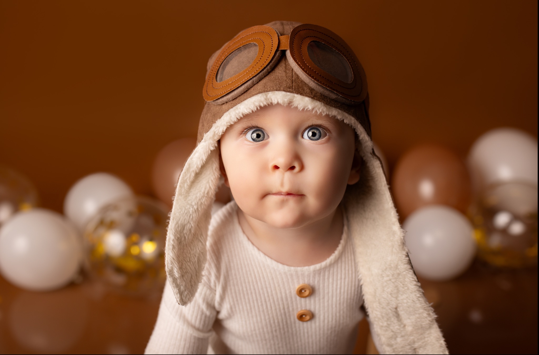 Portret niemowlęcia w brązowej czapce pilota z goglami, na rozmytym tle z balonami. Uwagę przykuwają duże, niebieskie oczy dziecka.