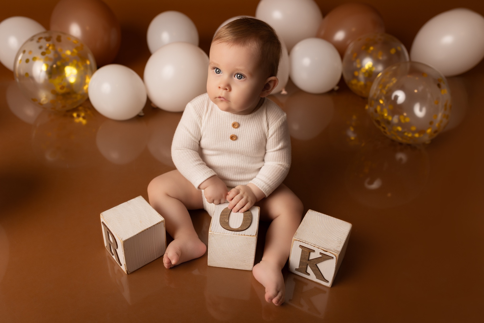 Urocze niemowlę w kremowym body siedzi na brązowym tle z białymi i złotymi balonami, otoczone drewnianymi klockami z literami. Dziecko patrzy w lewo.