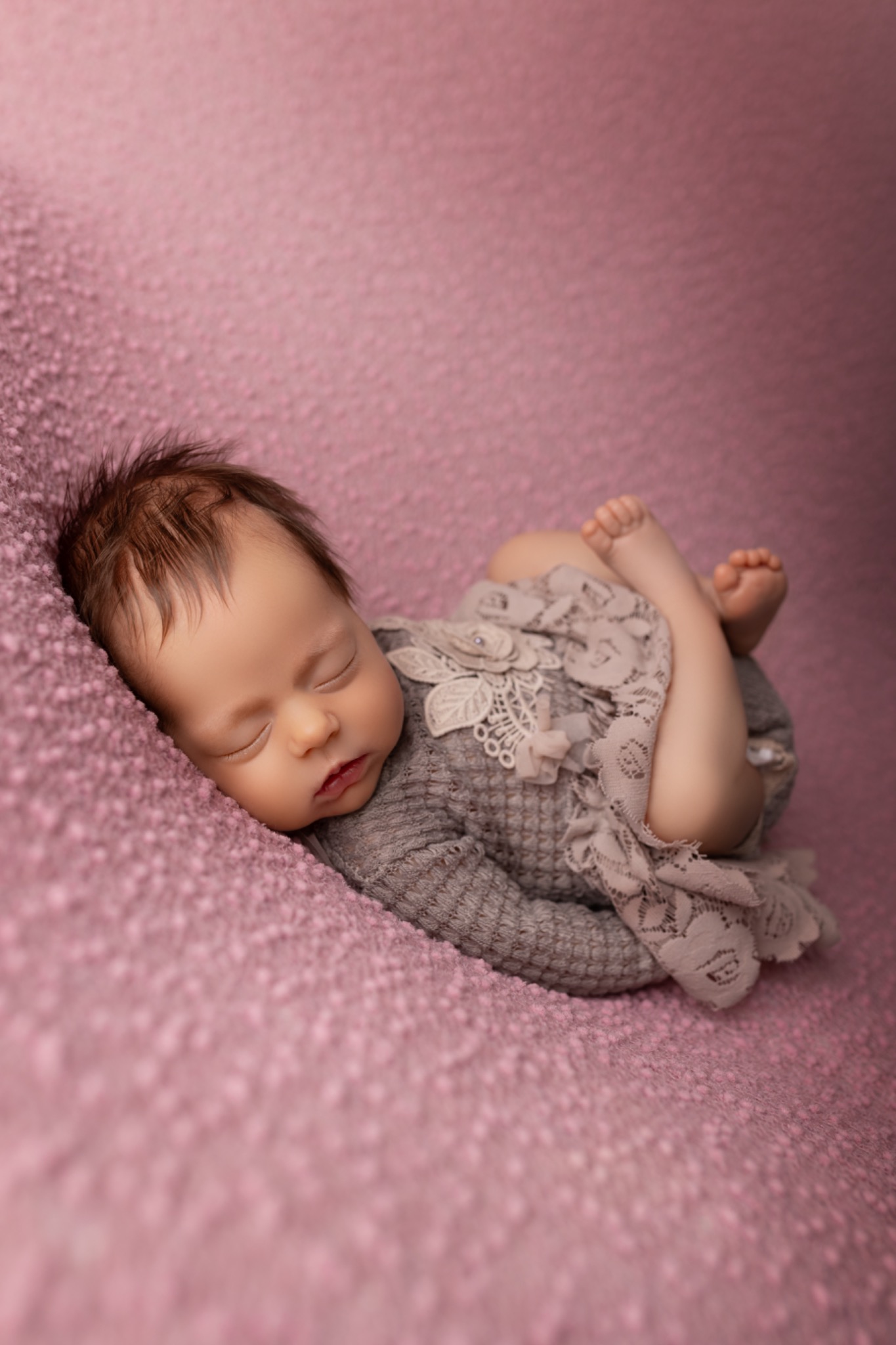 Śpiący noworodek w szarym ubranku z koronką, leżący na różowym, teksturowanym tle. Delikatne światło podkreśla rysy twarzy dziecka.