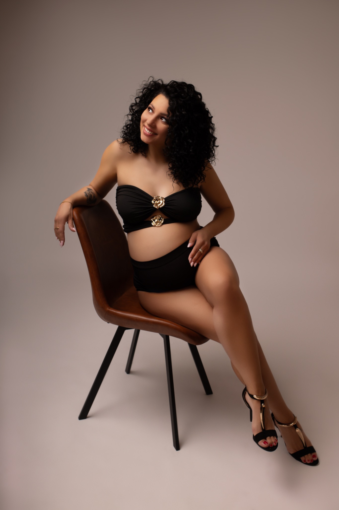 Kobieta w ciąży, w czarnym stroju kąpielowym, siedzi na brązowym krześle. Uśmiechnięta, patrzy w górę. Sesja w studio na jasnym tle. Eleganckie buty na obcasie.
