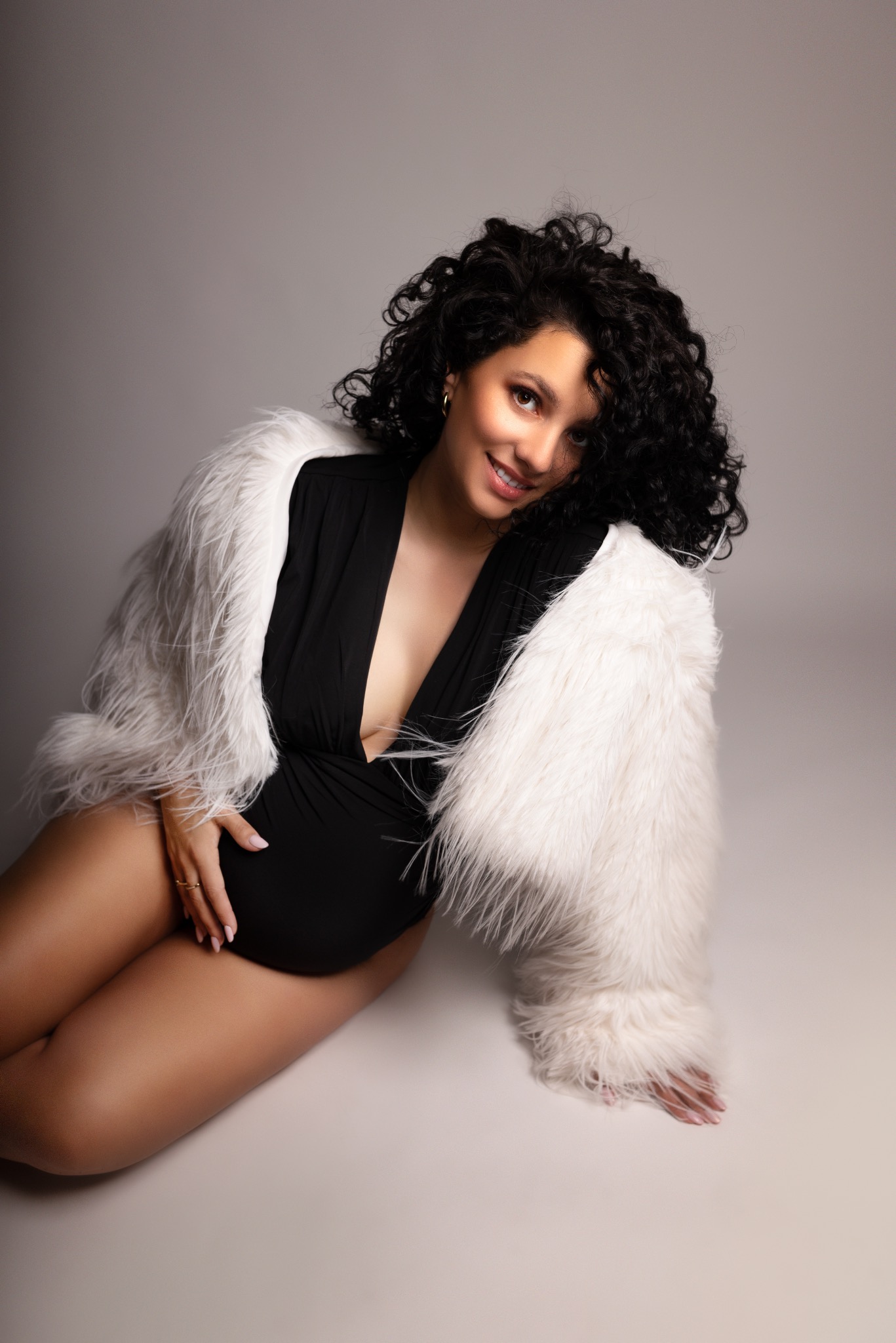 Uśmiechnięta, ciemnowłosa kobieta w czarnym body i białym futrze, leżąca na podłodze. Dłoń na brzuchu, subtelny makijaż, ciepłe oświetlenie.