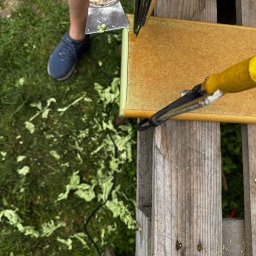 Michał Idzikowski - Usuwanie starej farby z płyty meblowej przy użyciu opalarki i szpachelki na tle trawy. Widoczne resztki farby. Praca na zewnątrz.