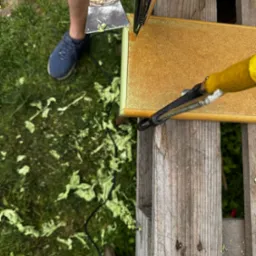 Usuwanie starej farby z płyty meblowej przy użyciu opalarki i szpachelki na tle trawy. Widoczne resztki farby. Praca na zewnątrz.