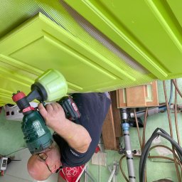 Michał Idzikowski - Mężczyzna maluje pistolem natryskowym zielone drzwi z frezowanymi wzorami. Częściowo pomalowane, widoczne niedomalowane fragmenty. Ujęcie z bliska, narzędzia pracy.