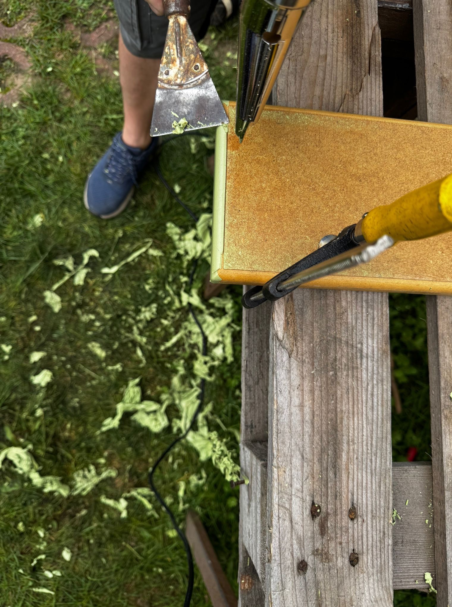 Usuwanie starej farby z płyty meblowej przy użyciu opalarki i szpachelki na tle trawy. Widoczne resztki farby. Praca na zewnątrz.