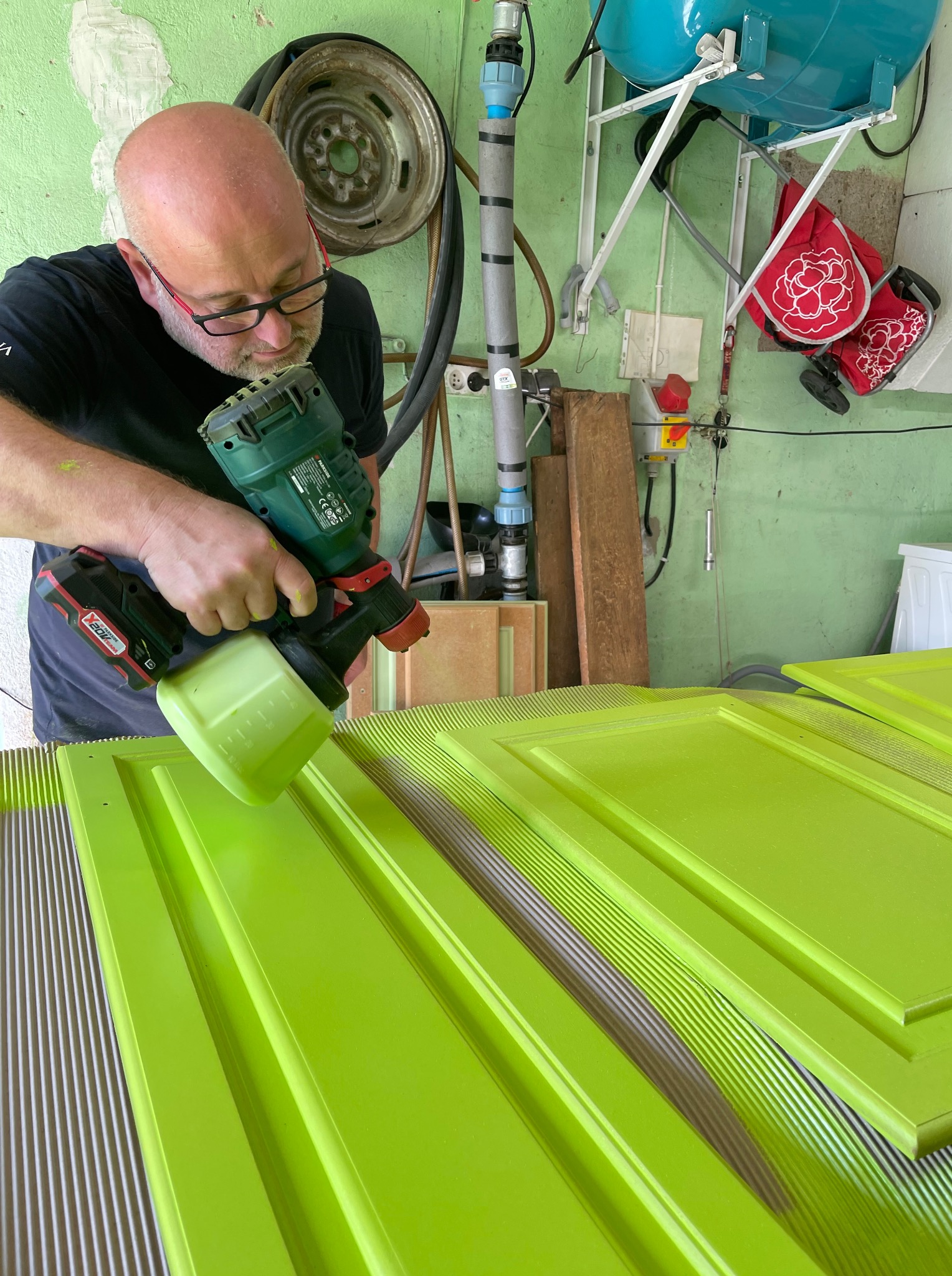 Mężczyzna w okularach maluje natryskowo zieloną farbą drzwi na stojaku. W tle zielona ściana, narzędzia i akcesoria. Używa elektrycznego pistoletu natryskowego.