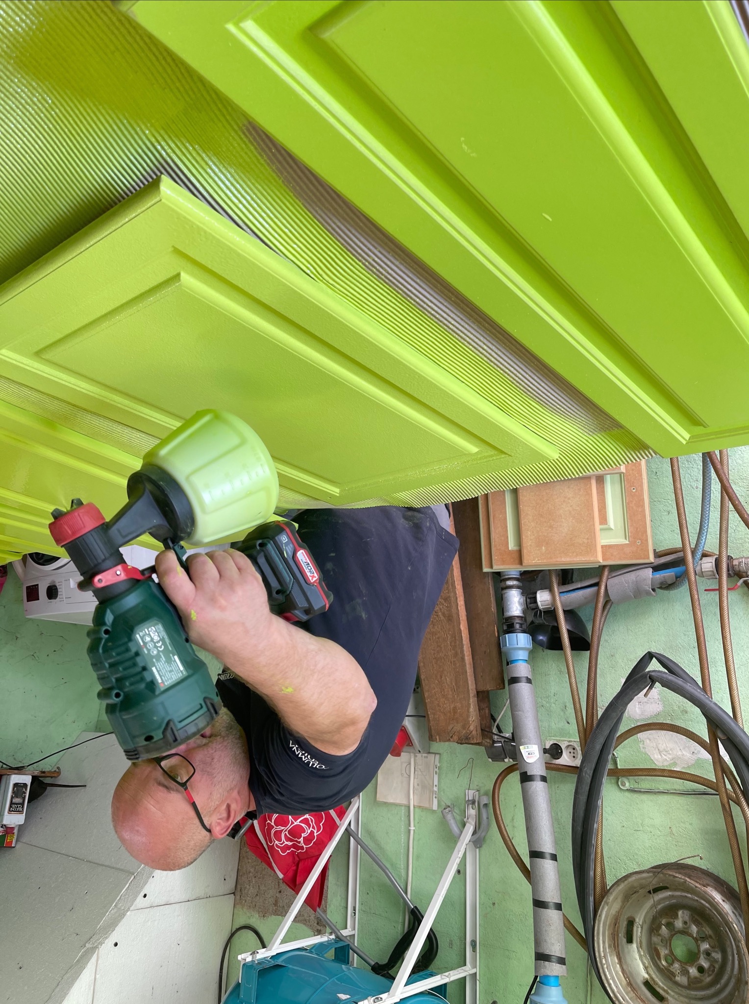 Mężczyzna maluje pistolem natryskowym zielone drzwi z frezowanymi wzorami. Częściowo pomalowane, widoczne niedomalowane fragmenty. Ujęcie z bliska, narzędzia pracy.