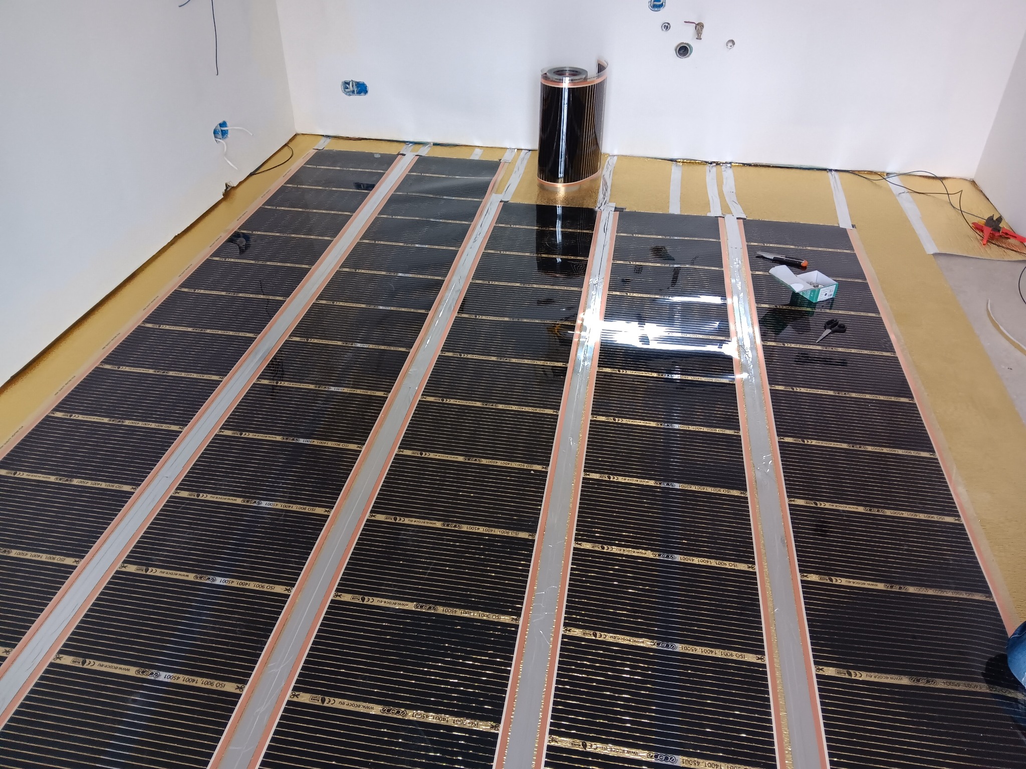 Montaż folii grzewczej na podłodze, widoczne narzędzia i rolka folii. Przygotowanie do dalszych prac wykończeniowych, instalacja ogrzewania podłogowego.