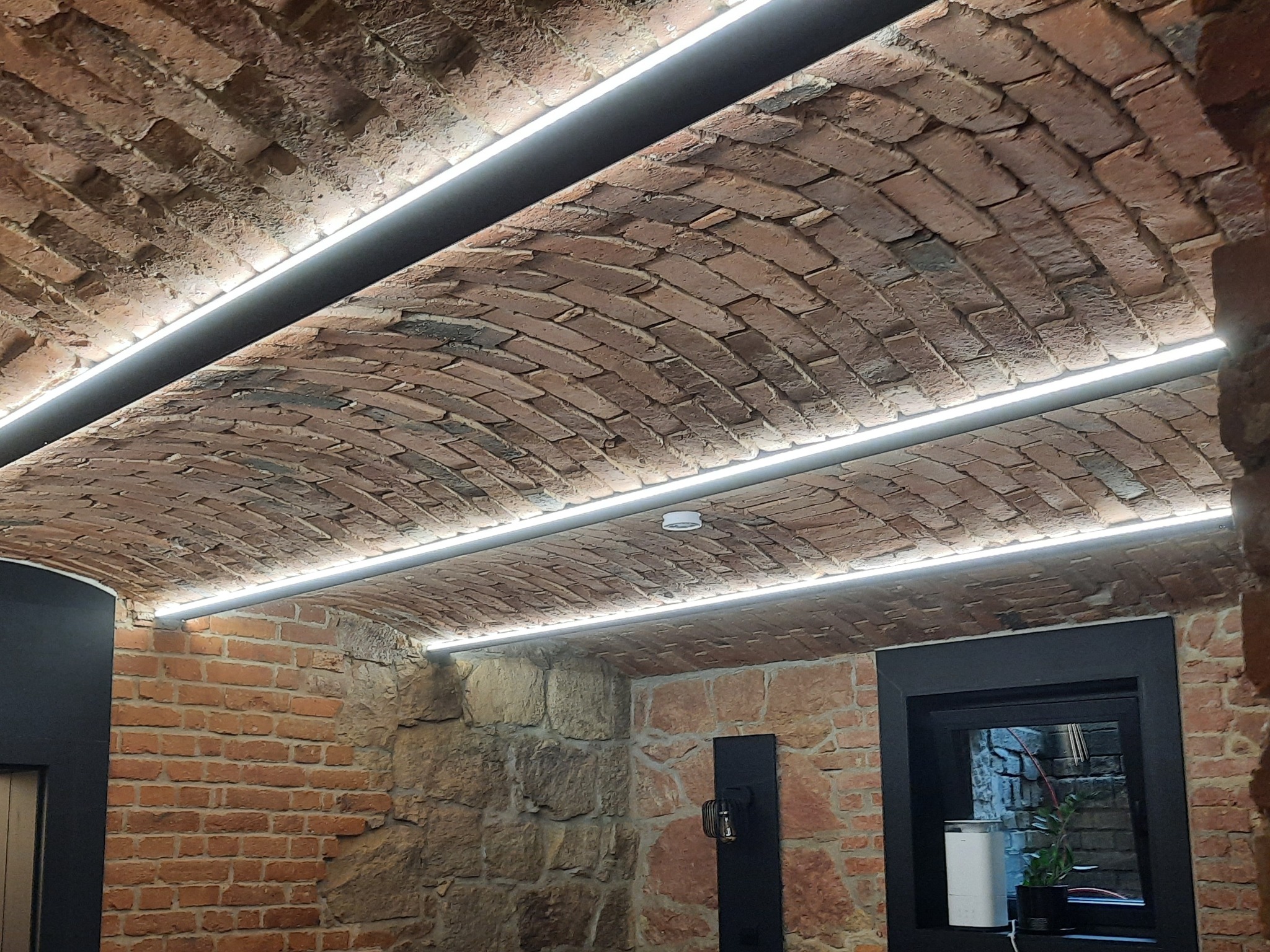 Ceglany łuk sufitowy z oświetleniem LED, czarna rama okienna, fragment kamiennej ściany. Nietypowe wykończenie wnętrza z akcentem na naturalne materiały i nowoczesne oświetlenie.