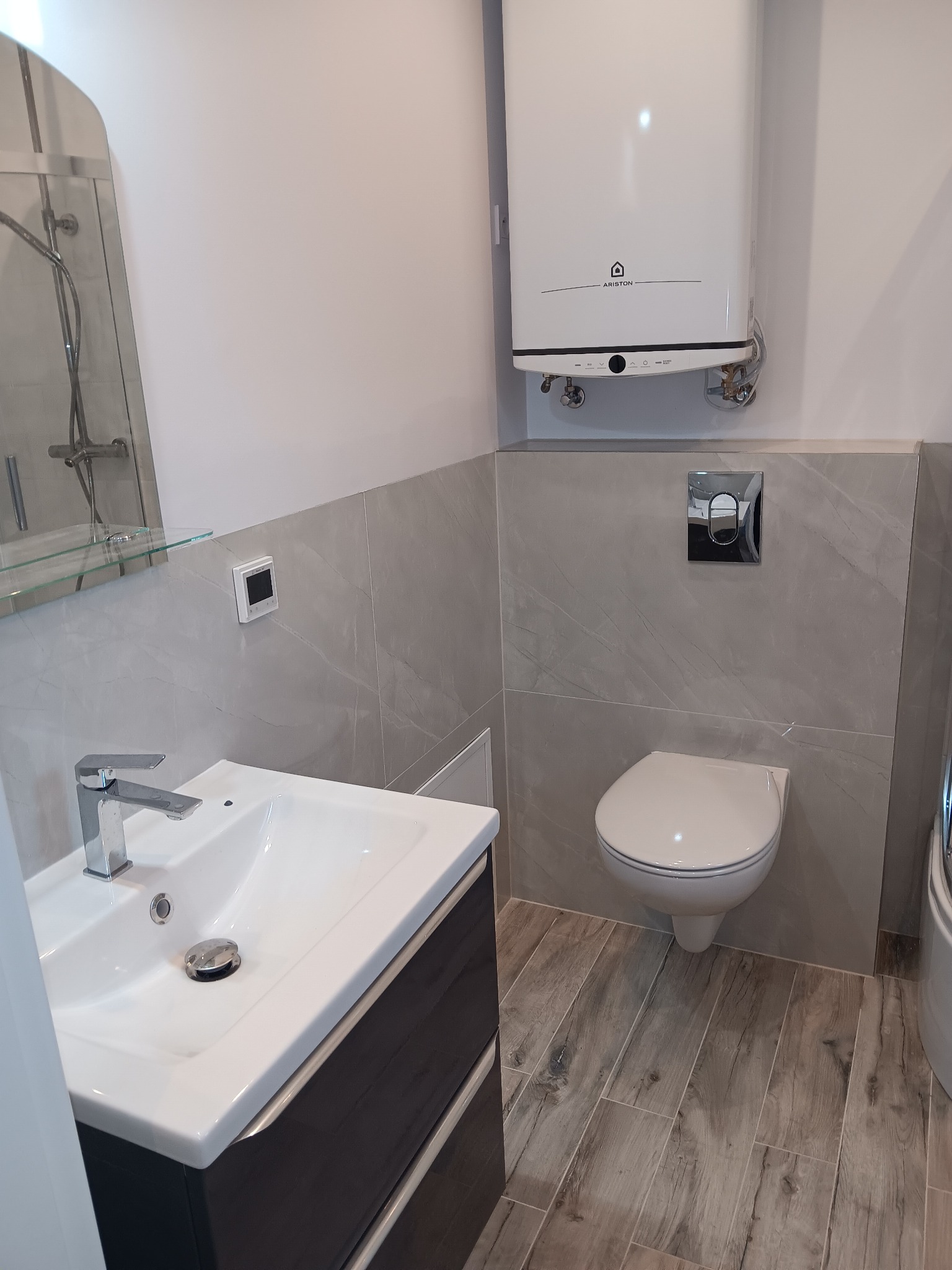 Wyremontowana łazienka z szarą glazurą imitującą marmur, drewnopodobną podłogą, umywalką z szafką i podgrzewaczem wody Ariston na ścianie.