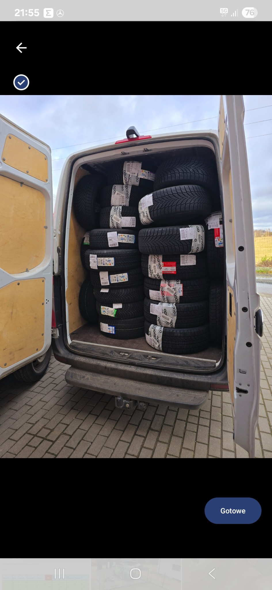 Wnętrze białego busa wypełnione oponami zimowymi, zabezpieczonymi taśmą z logo. Widoczne otwarte drzwi i fragment nawierzchni z kostki brukowej. Transport opon.