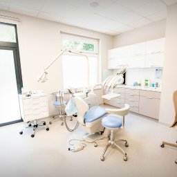 BUDKOWSKI DENTAL CLINIC - Jasne wnętrze gabinetu stomatologicznego z nowoczesnym fotelem, lampą i szafkami. Duże okno i drzwi w tle. Minimalistyczny design, brak pacjenta.