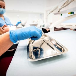 BUDKOWSKI DENTAL CLINIC - Stomatolog przygotowuje narzędzia na metalowej tacy podczas zabiegu. Pacjentka leży na fotelu, a lekarz w maseczce asystuje w tle.