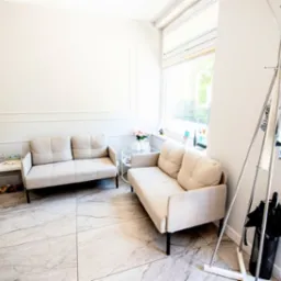 Jasne wnętrze poczekalni w Konstantynowie Łódzkim: dwie beżowe sofy, stolik z kwiatami, wieszak na ubrania i marmurowa podłoga. Przytulny design.