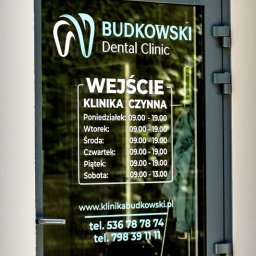 BUDKOWSKI DENTAL CLINIC - Szklane drzwi wejściowe do kliniki dentystycznej Budkowski Dental Clinic z logo i godzinami otwarcia. Widoczny adres strony i numery telefonów.