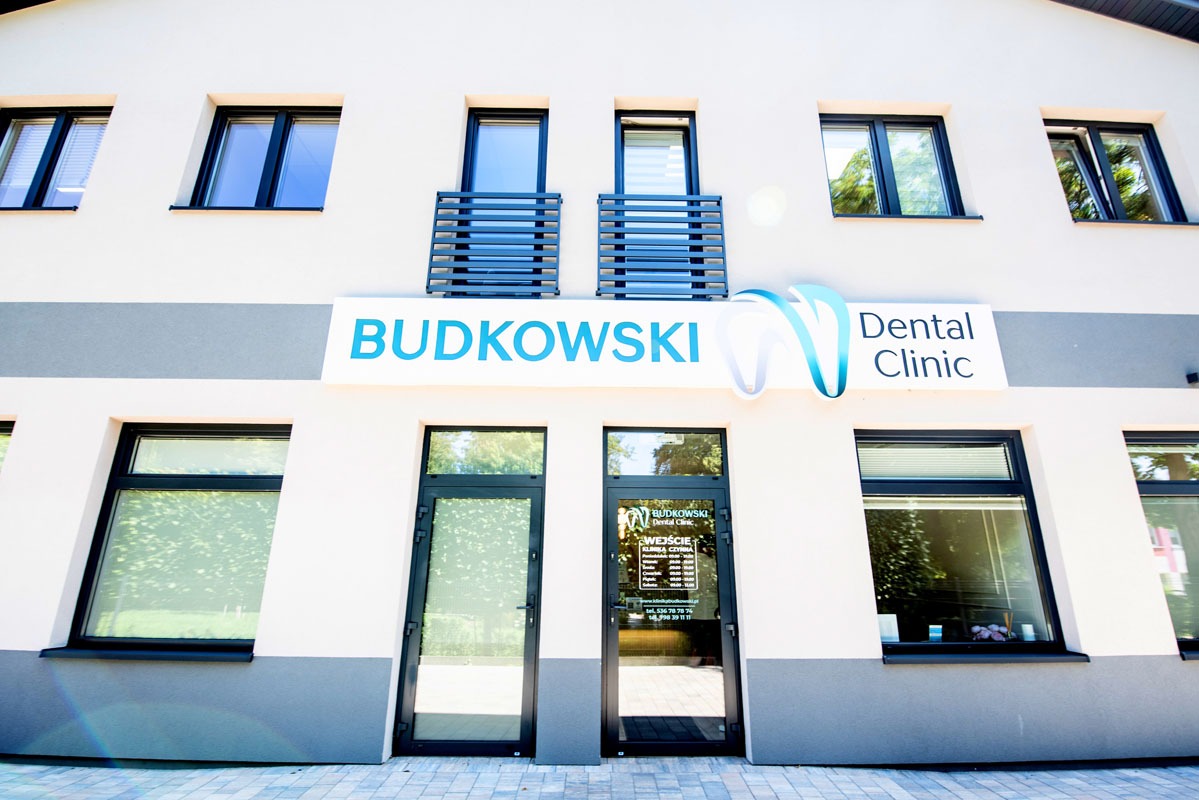 Elewacja budynku z szyldem Budkowski Dental Clinic. Jasna fasada z ciemnymi oknami i drzwiami wejściowymi. Nad wejściem logo kliniki i nazwa w kolorze niebieskim.