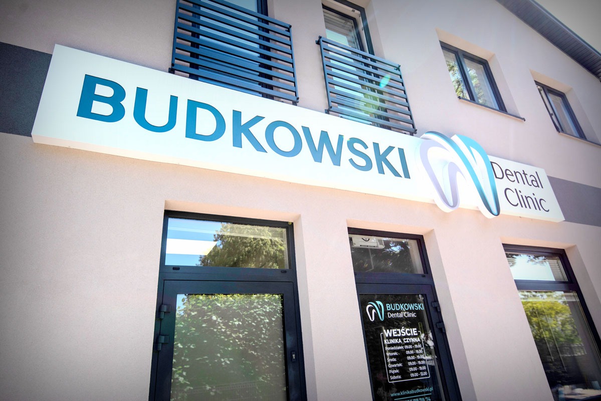 Fasada kliniki dentystycznej Budkowski z logo i godzinami otwarcia na drzwiach wejściowych, w słoneczny dzień. Balkony z metalowymi barierkami.