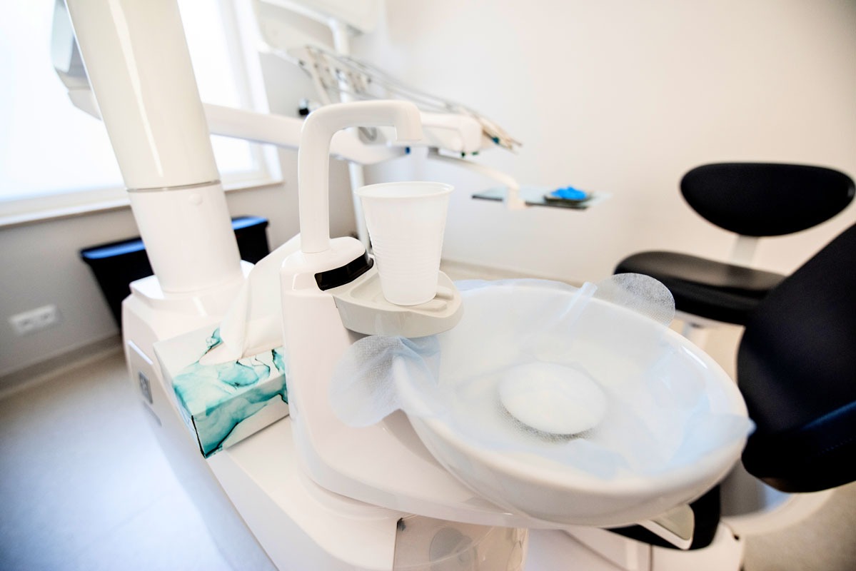 Nowoczesny unit stomatologiczny z białą misą, kubkiem i dozownikiem chusteczek w jasnym gabinecie. Widoczne narzędzia dentystyczne i fotel pacjenta w tle.