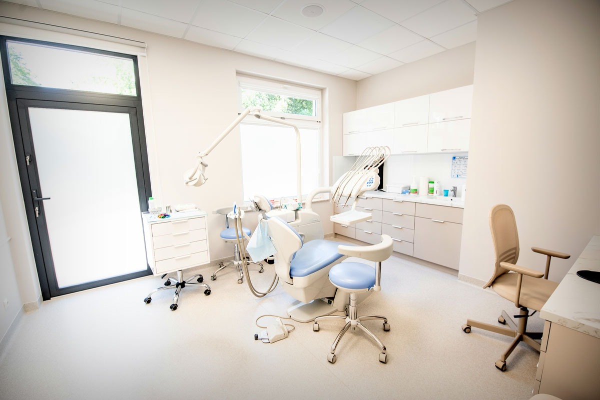 Jasne wnętrze gabinetu stomatologicznego z nowoczesnym fotelem, lampą i szafkami. Duże okno i drzwi w tle. Minimalistyczny design, brak pacjenta.