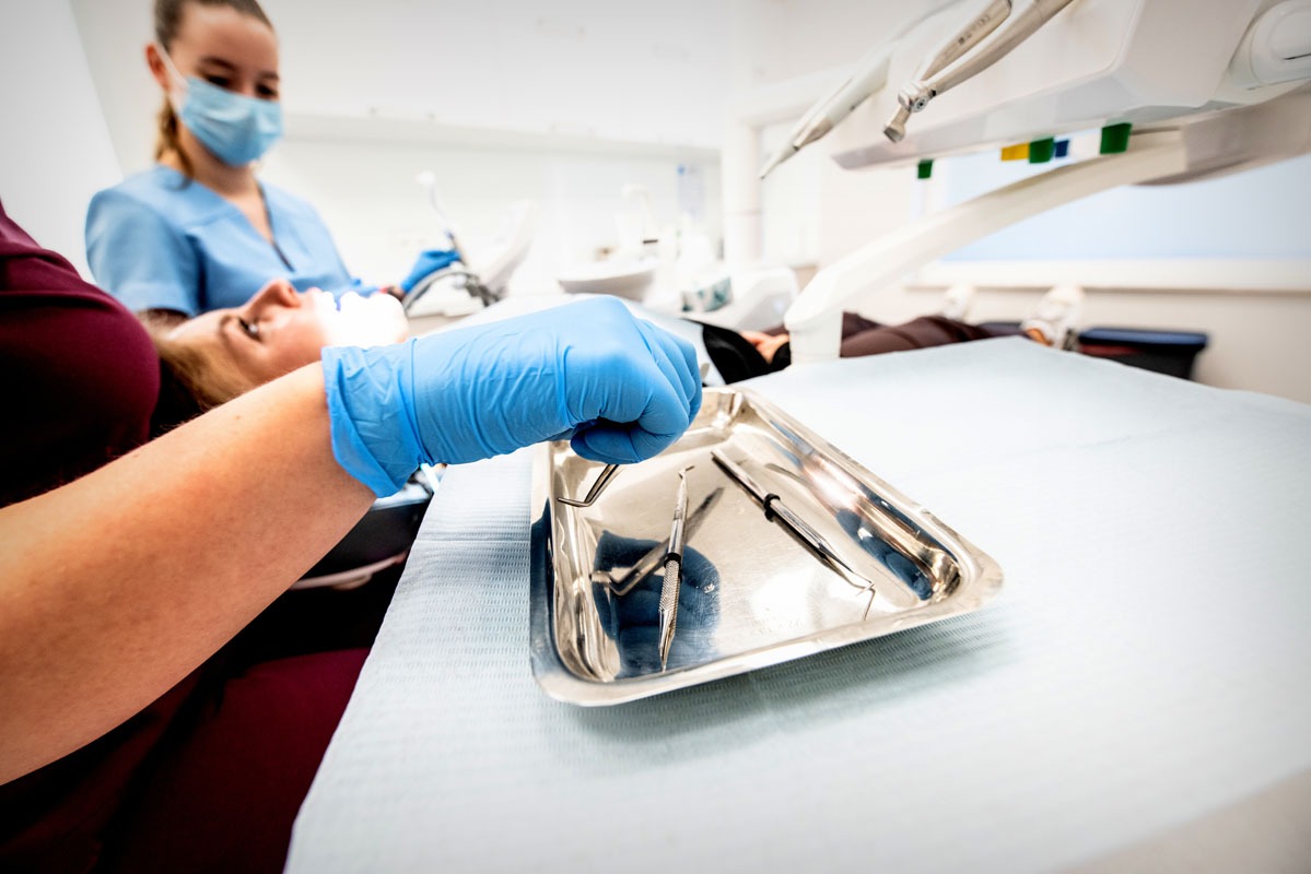 Stomatolog przygotowuje narzędzia na metalowej tacy podczas zabiegu. Pacjentka leży na fotelu, a lekarz w maseczce asystuje w tle.