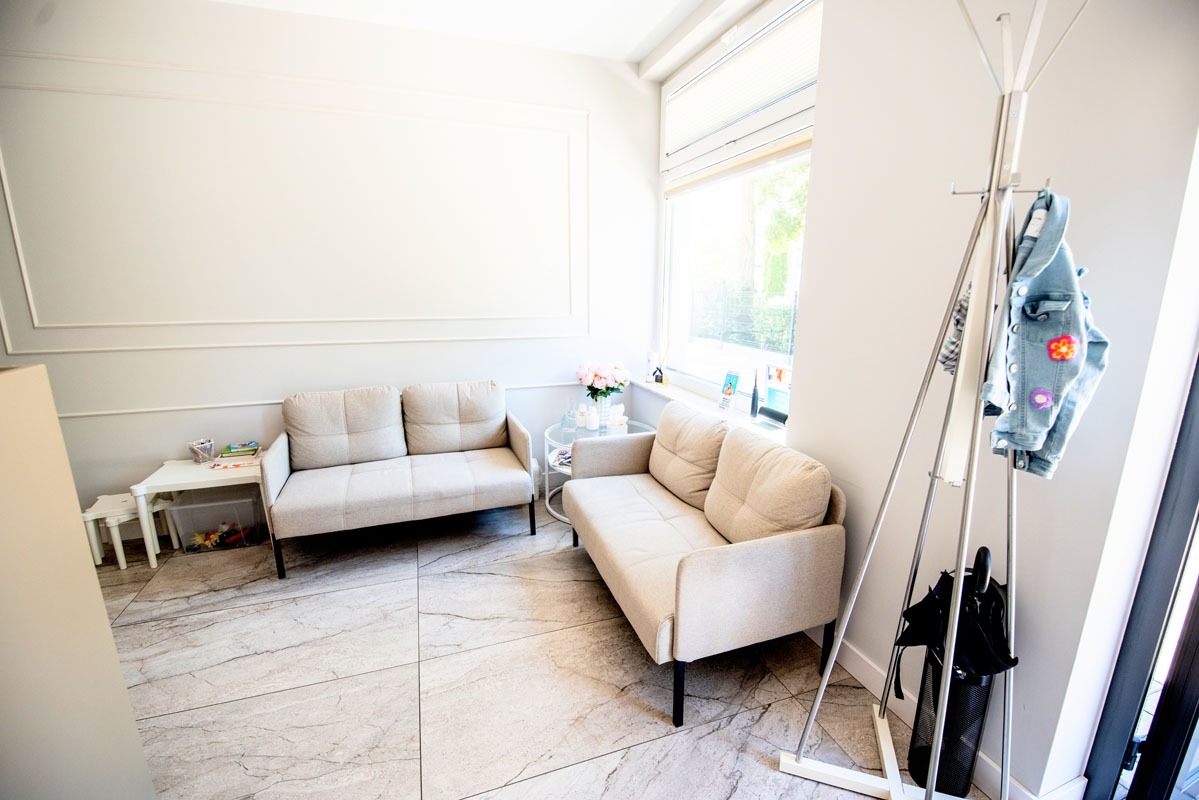 Jasne wnętrze poczekalni w Konstantynowie Łódzkim: dwie beżowe sofy, stolik z kwiatami, wieszak na ubrania i marmurowa podłoga. Przytulny design.