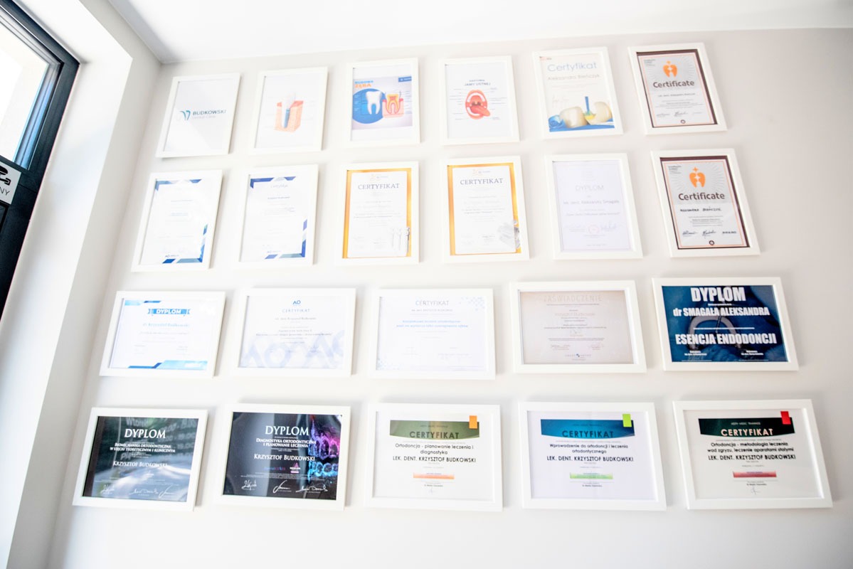 Ściana w klinice z ramkami zawierającymi dyplomy i certyfikaty lek. dent. Krzysztofa Budkowskiego i dr Smagały Aleksandry, z logo kliniki w górnym rzędzie.