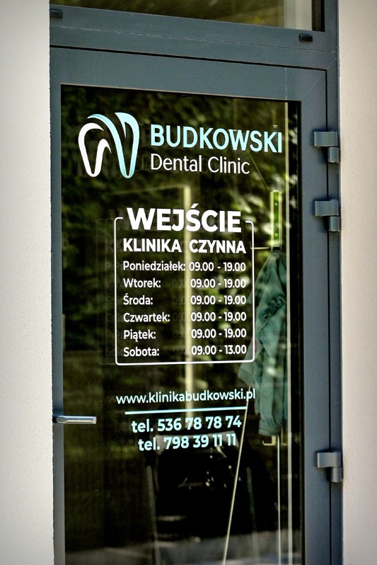 Szklane drzwi wejściowe do kliniki dentystycznej Budkowski Dental Clinic z logo i godzinami otwarcia. Widoczny adres strony i numery telefonów.