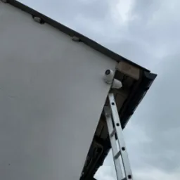 Dwie kamery monitoringu zamontowane na elewacji budynku, widoczne z dołu, z drabiną opartą o ścianę. Pochmurne niebo w tle.