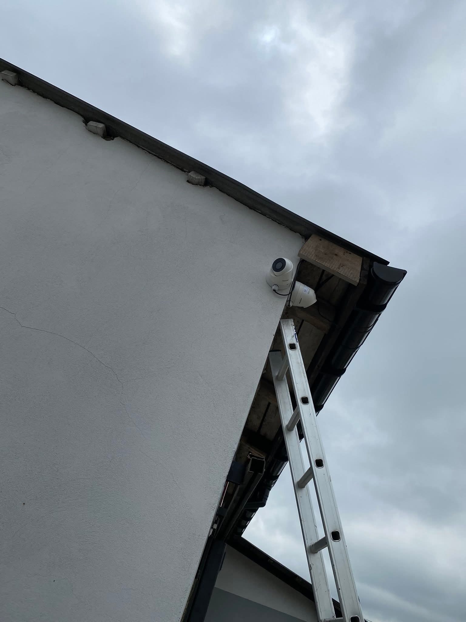 Dwie kamery monitoringu zamontowane na elewacji budynku, widoczne z dołu, z drabiną opartą o ścianę. Pochmurne niebo w tle.