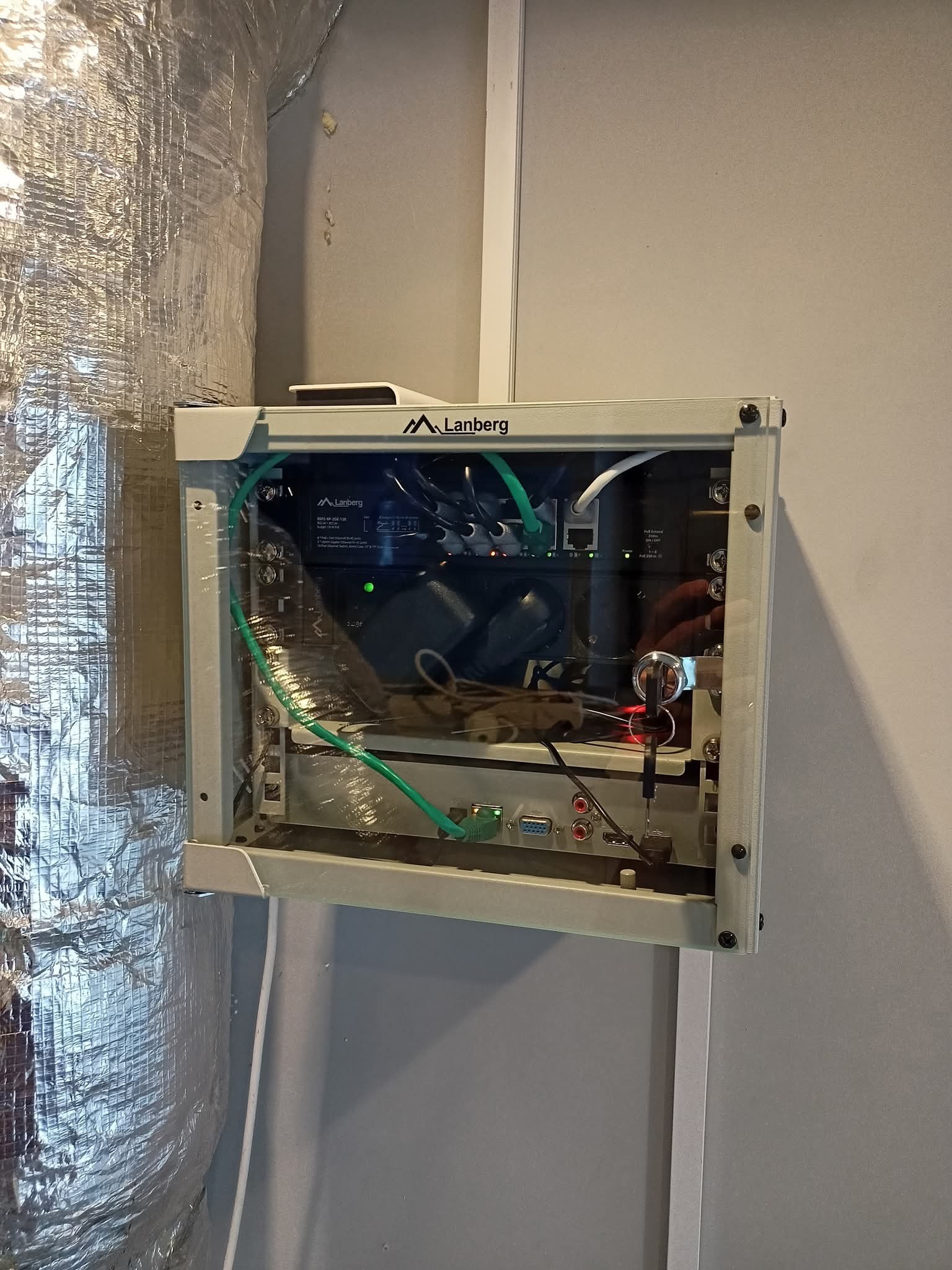 Zamknięta szafka Lanberg z widocznymi kablami i urządzeniami, zamontowana na szarej ścianie obok izolowanego przewodu wentylacyjnego.