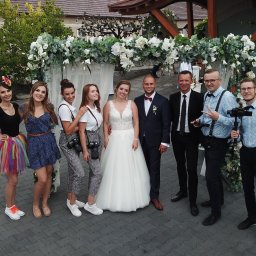 MagMar Studio Łukasz Kapral - Uśmiechnięta para młoda pozuje z zespołem fotografów i filmowców pod kwiatową bramą. Widoczni kamerzyści z profesjonalnym sprzętem i osoby w barwnych strojach.