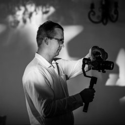 MagMar Studio Łukasz Kapral - Czarno-białe zdjęcie kamerzysty z gimbalem i kamerą, profil, oświetlenie studyjne, cień na ścianie, elegancki żyrandol w tle, przygotowanie do filmowania.