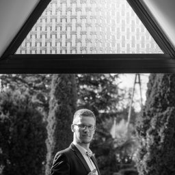MagMar Studio Łukasz Kapral - Czarno-białe zdjęcie mężczyzny w garniturze, trzymającego aparat, z trójkątnym oknem i zielenią w tle. Ujęcie z dołu, podkreślające architekturę i postać.