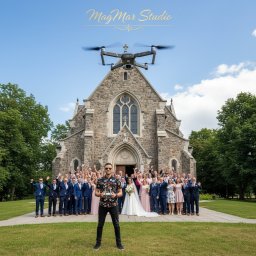 MagMar Studio Łukasz Kapral - Dron nad parą młodą i gośćmi weselnymi przed kamiennym kościołem, pilot drona w pierwszym planie. Ujęcie z drona na tle błękitnego nieba.