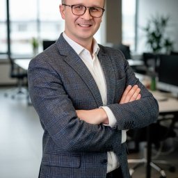 MagMar Studio Łukasz Kapral - Portret mężczyzny w okularach i marynarce w biurowym otoczeniu, z założonymi rękami i uśmiechem, na tle okien i biurek.