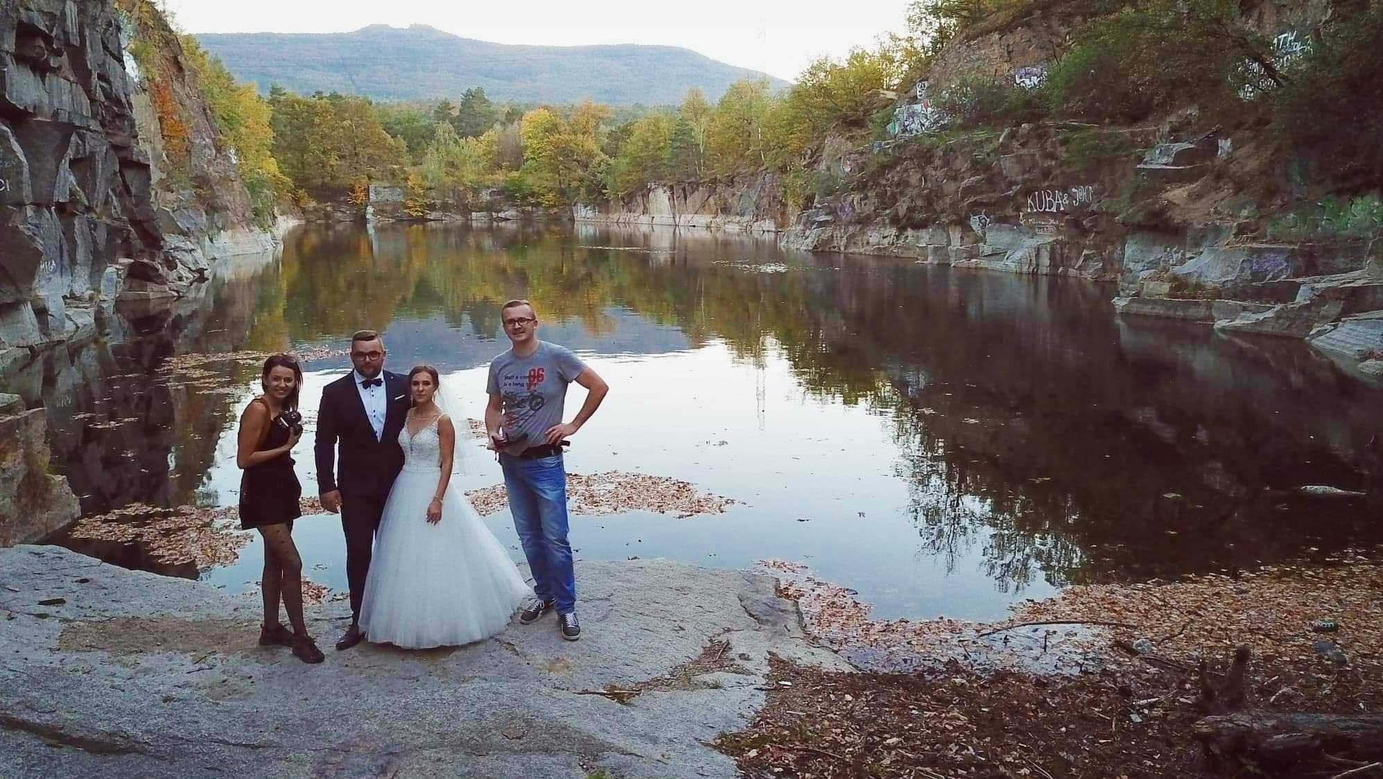Sesja ślubna w plenerze: para młoda, fotografka i asystent pozują na tle malowniczego jeziora w kamieniołomie, jesienne liście na wodzie, góry w oddali.