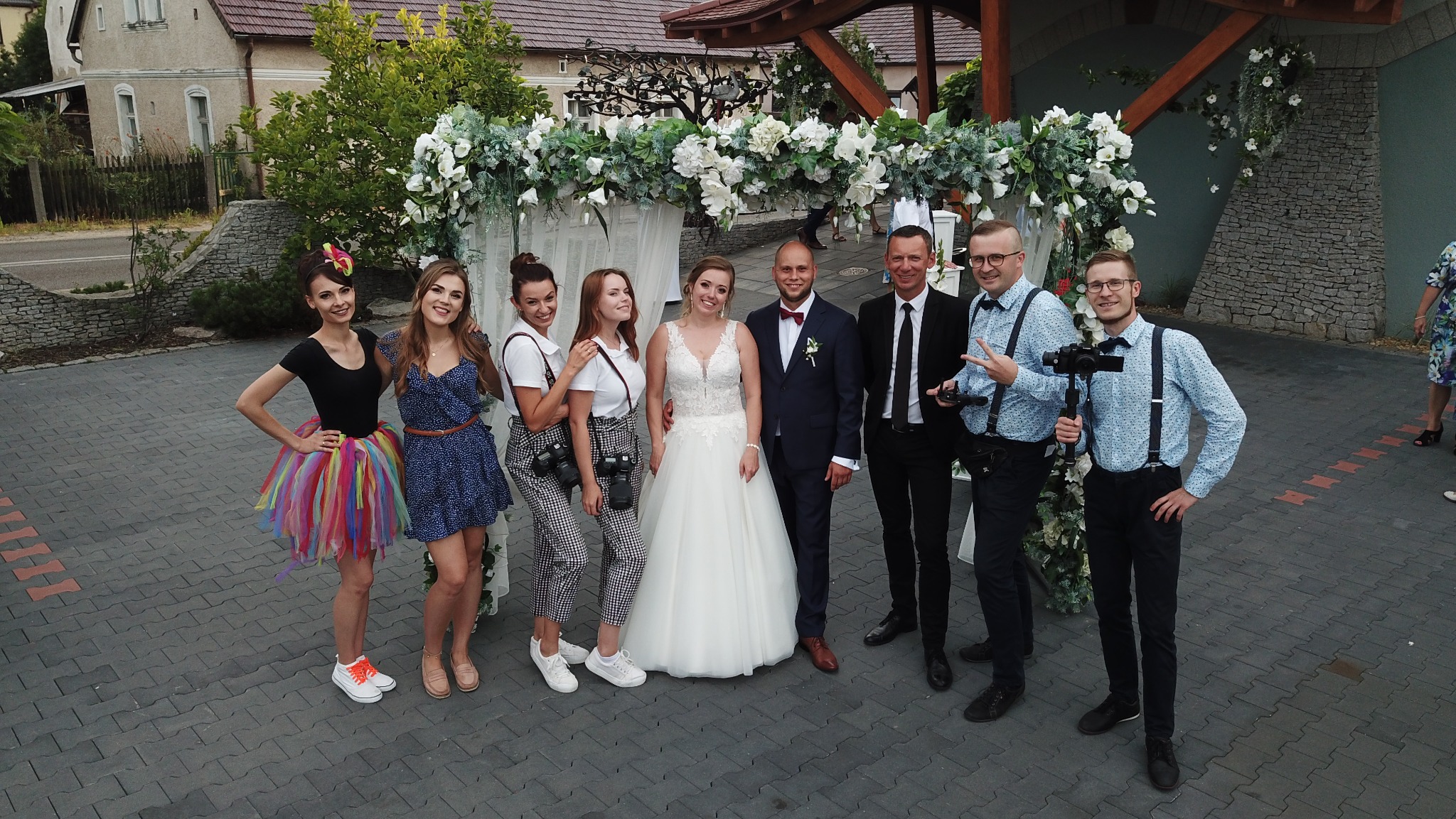 Uśmiechnięta para młoda pozuje z zespołem fotografów i filmowców pod kwiatową bramą. Widoczni kamerzyści z profesjonalnym sprzętem i osoby w barwnych strojach.