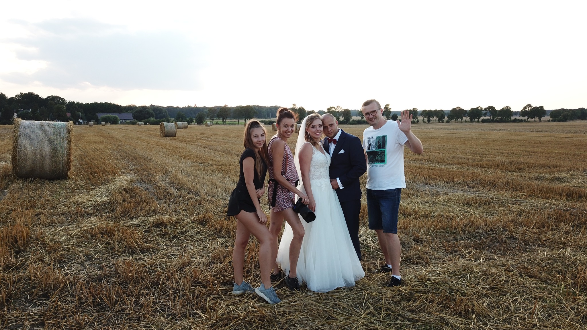 Sesja ślubna na polu ze słomą: para młoda w otoczeniu gości, fotograf z aparatem. Uśmiechy, naturalne światło, wiejski krajobraz w tle.