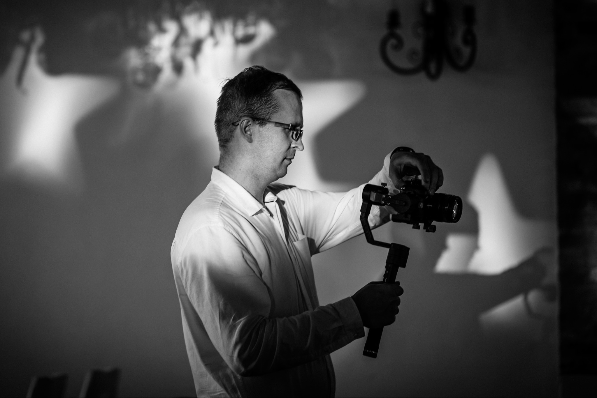 Czarno-białe zdjęcie kamerzysty z gimbalem i kamerą, profil, oświetlenie studyjne, cień na ścianie, elegancki żyrandol w tle, przygotowanie do filmowania.