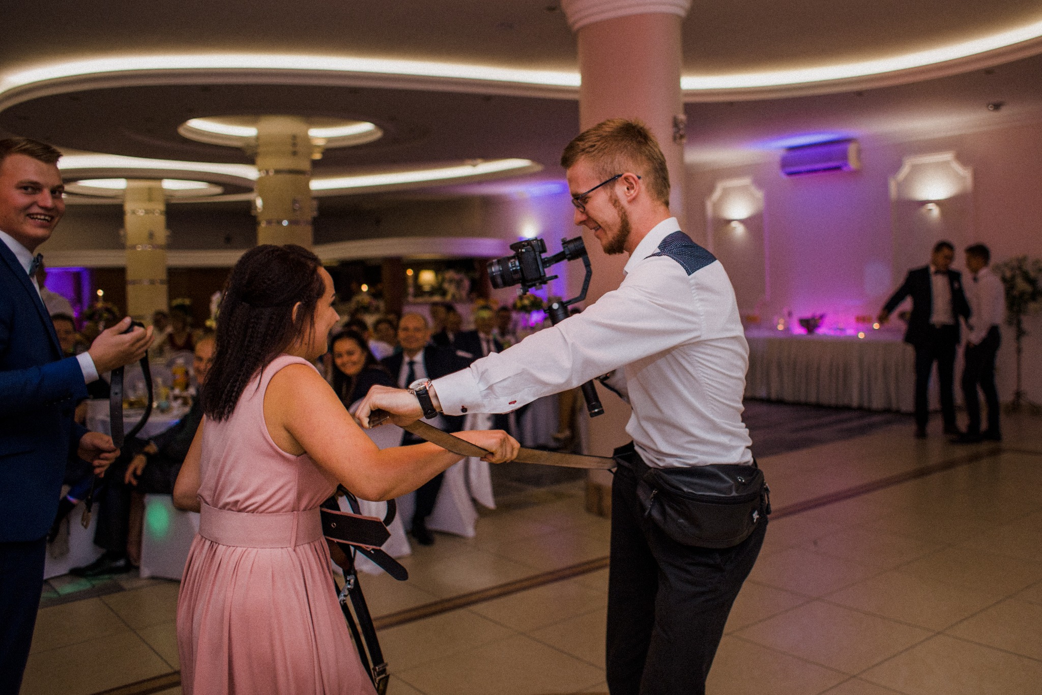 Kamerzysta z gimbalem filmuje taniec z rekwizytami podczas wesela. W tle goście i dekoracje sali. Mężczyzna w okularach, biała koszula i czarne spodnie.