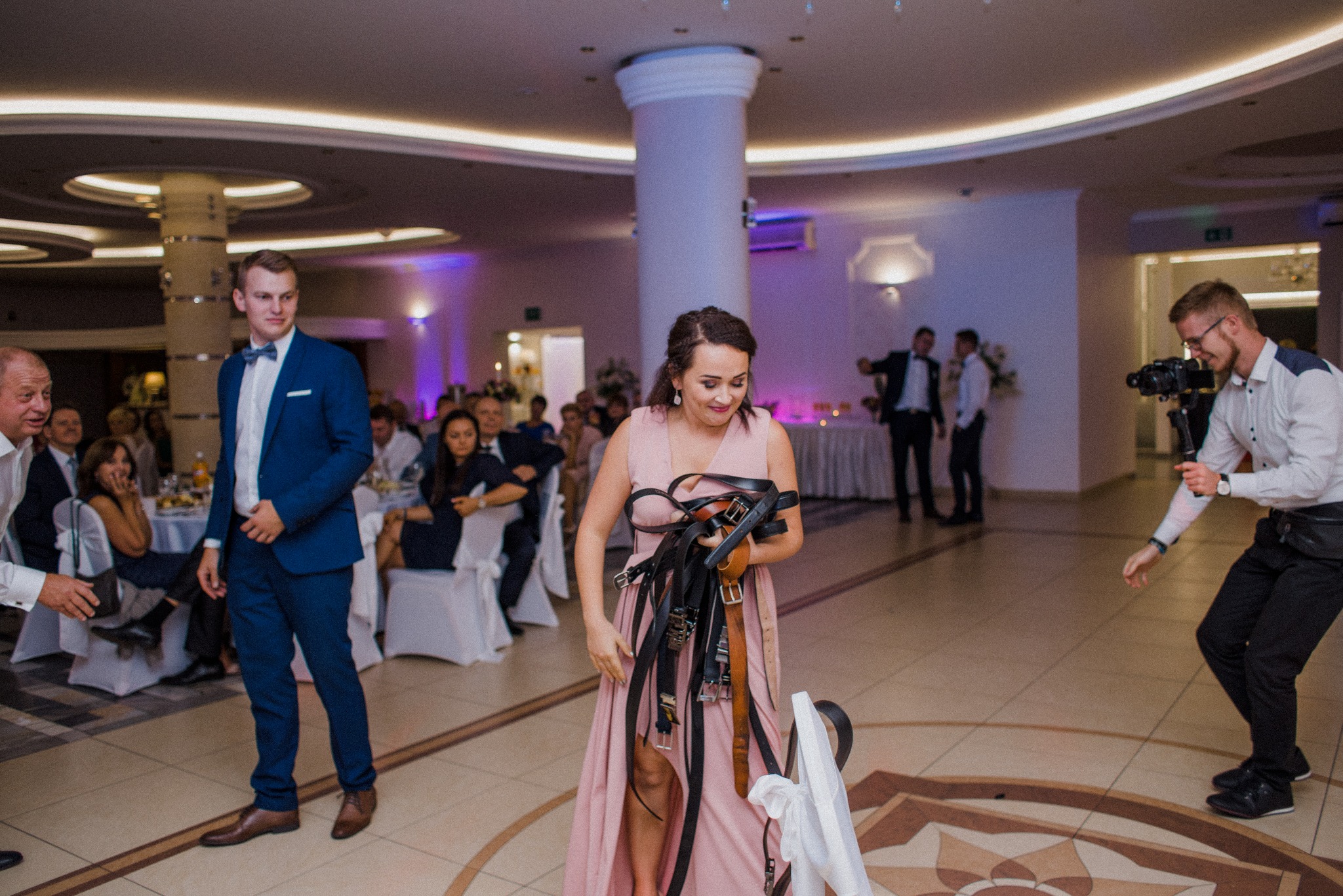 Kamerzysta filmuje kobietę z rekwizytami na sali balowej. W tle goście weselni i mężczyzna w niebieskim garniturze. Dekoracyjne kolumny i oświetlenie.