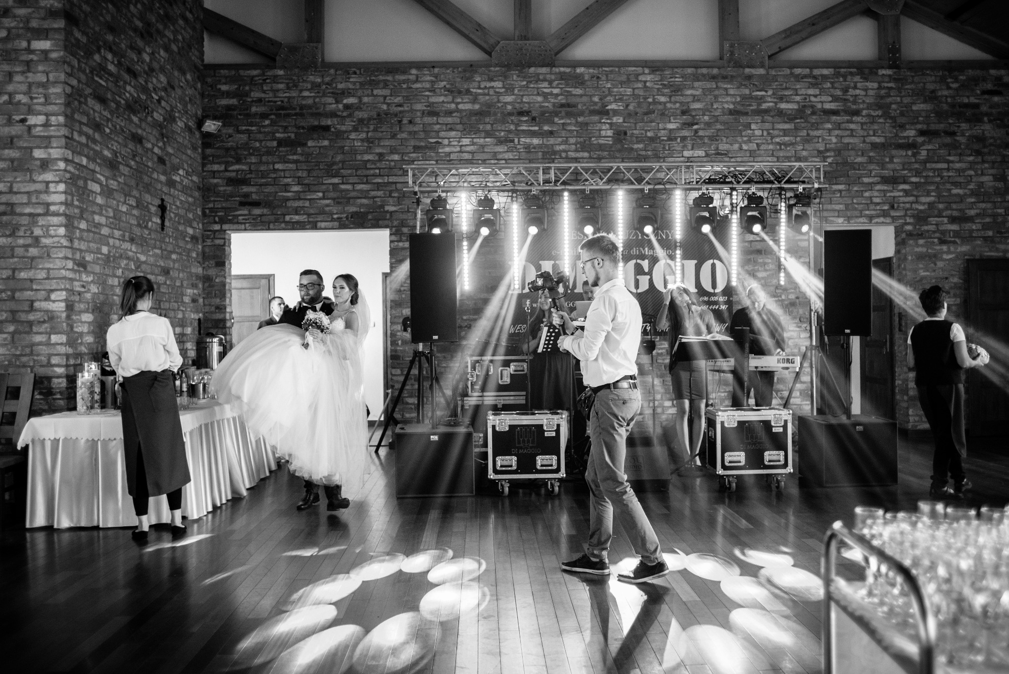 Czarno-białe zdjęcie kamerzysty filmującego parę młodą na sali weselnej z ceglanymi ścianami i oświetleniem scenicznym. Widoczny zespół muzyczny w tle.
