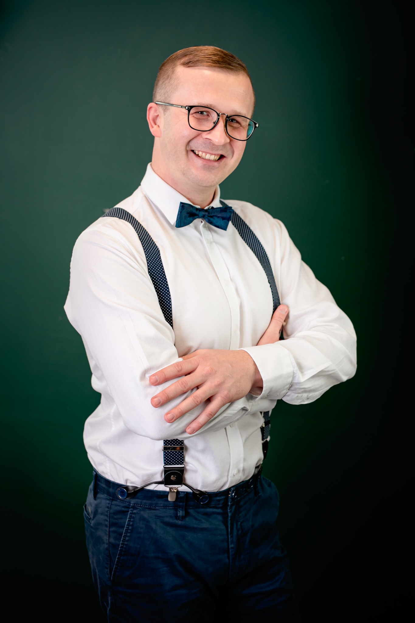 Uśmiechnięty mężczyzna w okularach, białej koszuli, muszce i szelkach na tle ciemnozielonym. Skrzyżowane ręce, elegancki strój, portret w studiu.