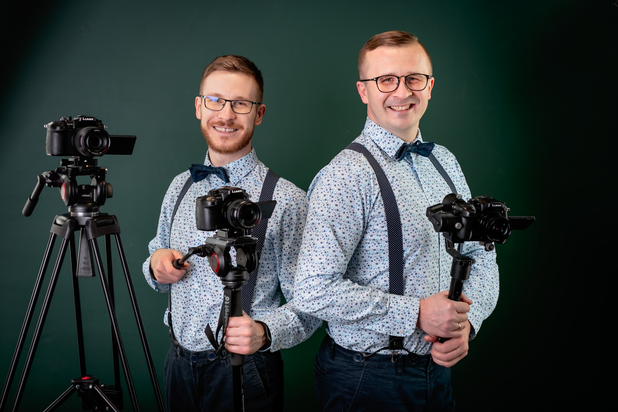 Dwóch uśmiechniętych mężczyzn w koszulach i muchach, prezentujących kamery Lumix na statywach i gimbalach na tle ciemnozielonego studia. Profesjonalny sprzęt.