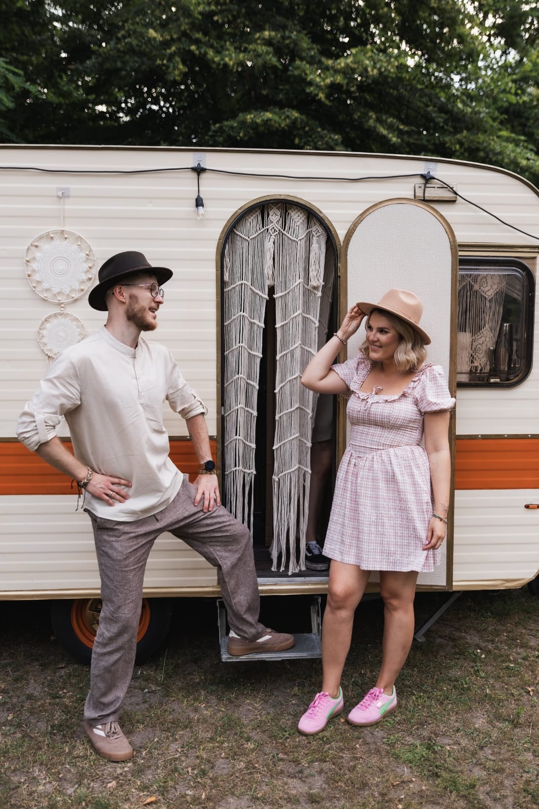Sesja narzeczeńska w stylu boho: para w kapeluszach pozuje przy kremowej przyczepie kempingowej z makramą w drzwiach. Uśmiechnięta kobieta poprawia kapelusz.