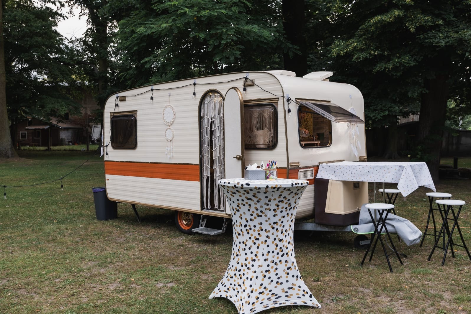 Klimatyczna fotobudka w stylu retro, umieszczona w przyczepie kempingowej z dekoracjami boho i stolikiem z akcesoriami na tle zieleni. Oświetlenie żarówkowe.