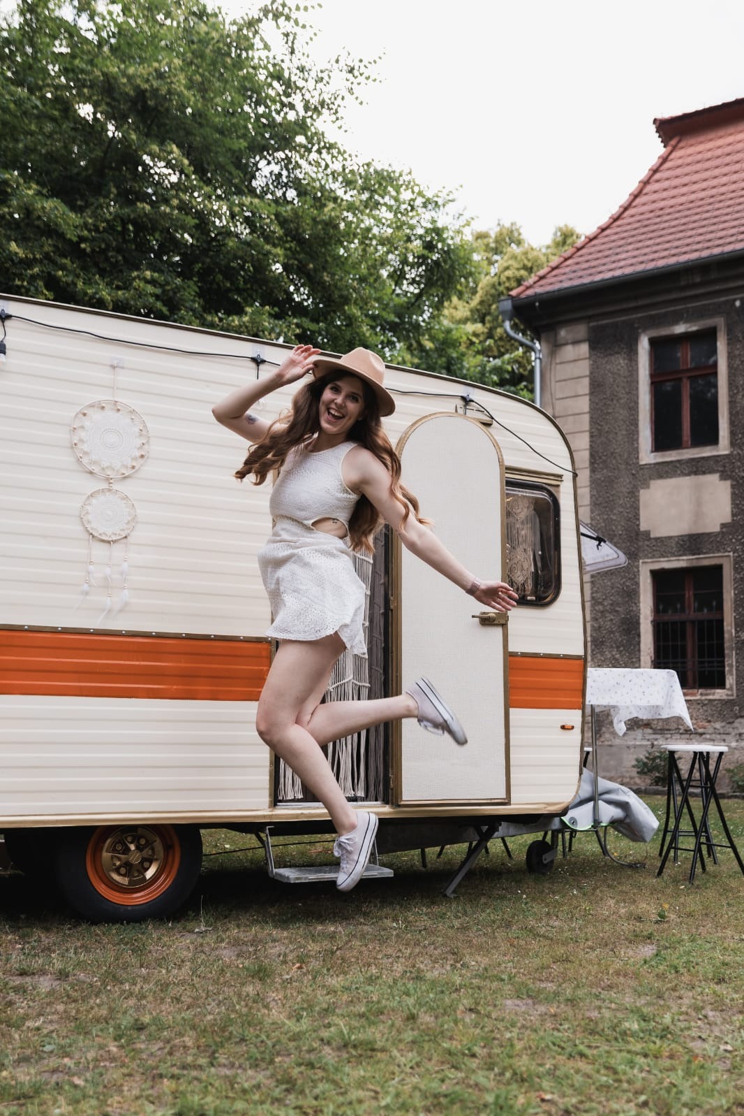 Uśmiechnięta kobieta w kapeluszu skacze przed kremową przyczepą kempingową z pomarańczowym pasem, na tle zieleni i budynku z czerwonym dachem.