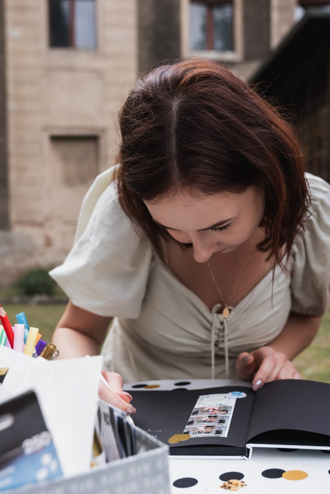 Kobieta w jasnej sukience wkleja pasek zdjęć z fotobudki do czarnego albumu, na stole pudełko z pisakami i konfetti. Tło stanowi budynek.