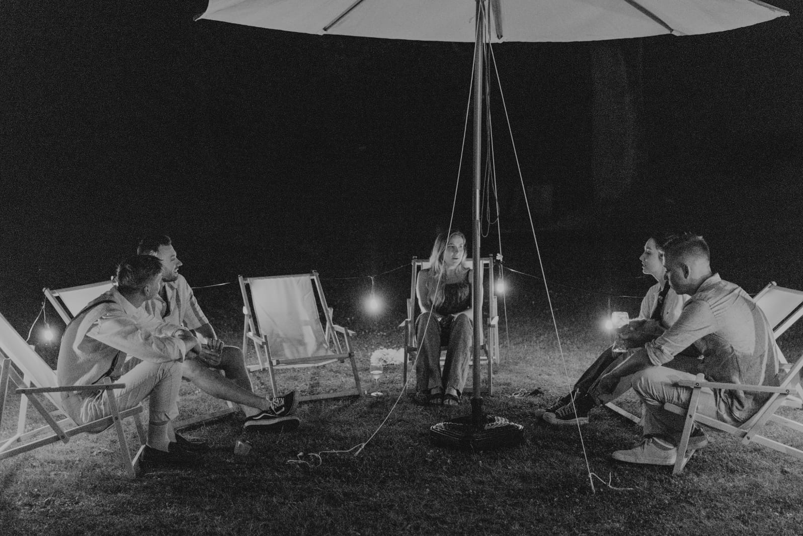 Czarno-białe zdjęcie grupy osób siedzących na leżakach pod parasolem w nocy, oświetlonych lampkami, tworzących intymną atmosferę spotkania.