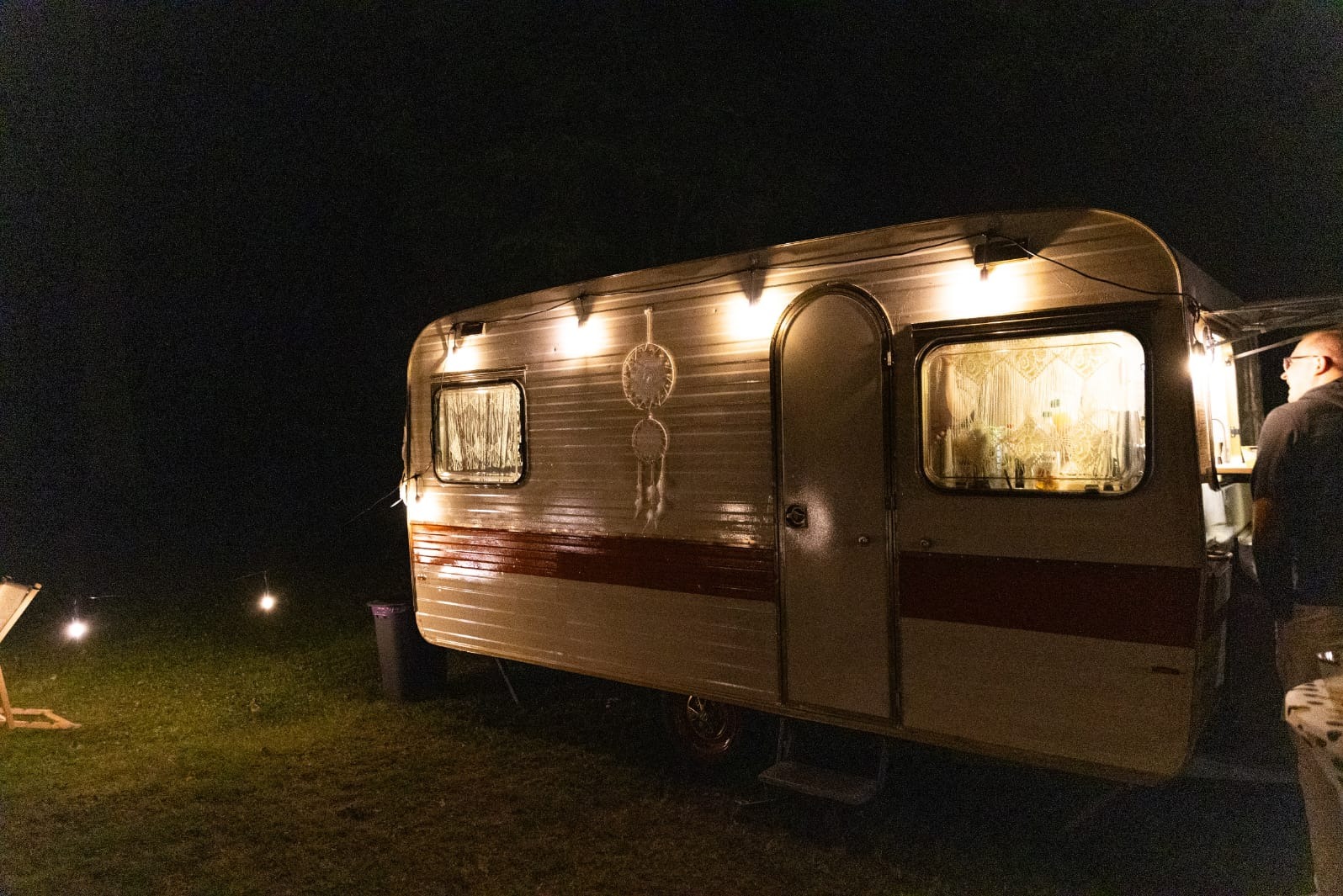 Oświetlona przyczepa kempingowa w nocy, z dekoracją łapacza snów i widocznym wnętrzem przez okno. Obok stoi mężczyzna, a w tle widać oświetlone krzesło.